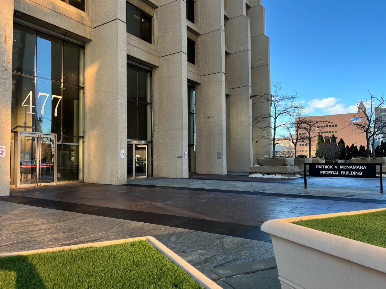 The Detroit Passport Agency. It is a tall concrete office building with a grid of windows. “477” is visible in large numbers above the front door. A sign near the entrance says “PATRICK V. McNAMARA FEDERAL BUILDING”.